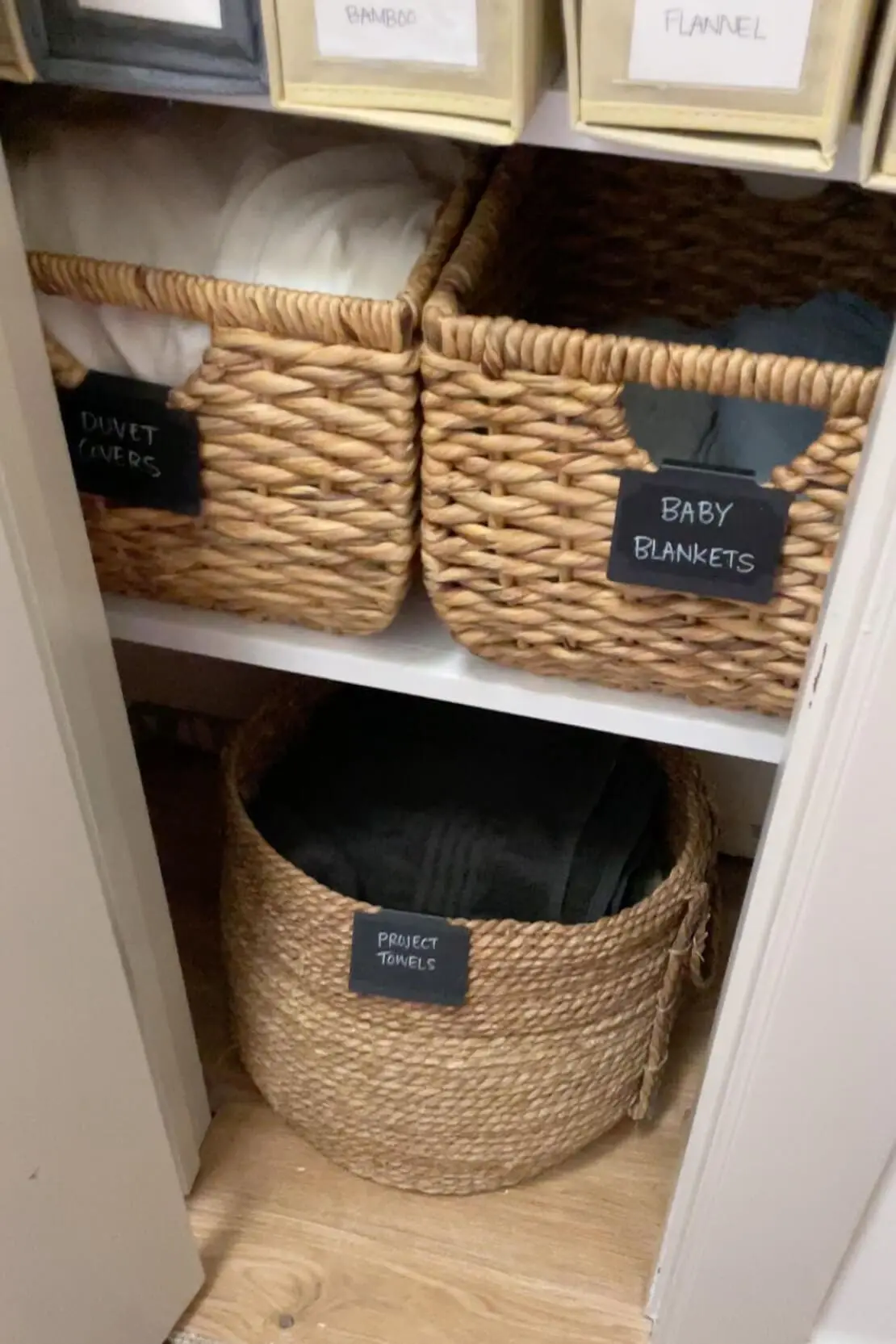 Linen closet storage featuring labeled woven baskets for baby blankets, duvet covers, and project towels.